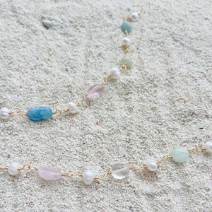 May include: A delicate necklace featuring a gold chain adorned with a variety of gemstones and pearls. The necklace includes light blue, pink, and clear stones, along with white pearls. The jewelry is set against a sandy background.