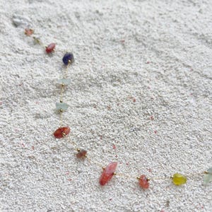 May include: A delicate gold chain necklace featuring a series of colorful, irregularly shaped gemstones. The gemstones are in shades of red, pink, green, yellow, and purple. The necklace is displayed on a bed of white sand.