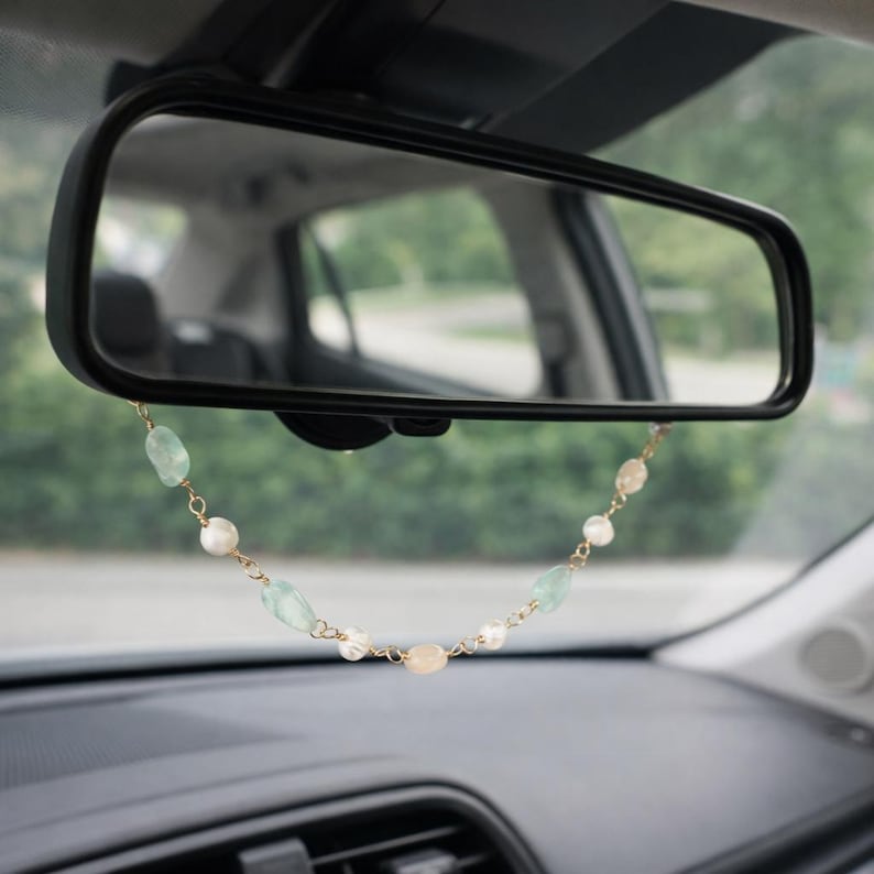 May include: A car rearview mirror with a decorative beaded chain hanging from it. The chain features a mix of pale green, white, and peach-colored beads strung on a gold-colored chain. The car's interior is visible in the background.