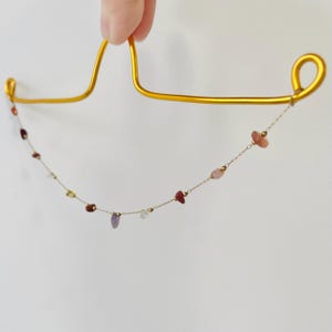 May include: A delicate gold necklace with a chain of small, colorful gemstones. The gemstones are various shapes and colors, including red, pink, purple, and yellow. The necklace is held up by a gold-colored hanger.
