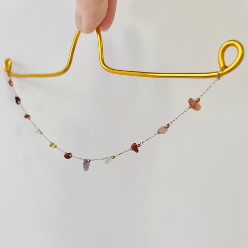 May include: A delicate gold necklace with a chain of small, colorful gemstones. The gemstones are various shapes and colors, including red, pink, purple, and yellow. The necklace is held up by a gold-colored hanger.