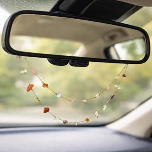 以下が含まれることがあります： 車のバックミラーから吊り下げられた、カラフルな宝石ビーズが付いたゴールドカラーのチェーン。 宝石はオレンジ、ピンク、クリアなど、さまざまな形と色をしています。 車内が背景に見えます。