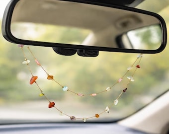 May include: A gold-colored chain with colorful gemstone beads hangs from a car's rearview mirror. The gemstones are various shapes and colors, including orange, pink, and clear. The car's interior is visible in the background.