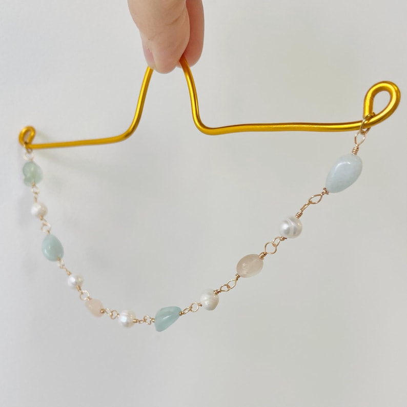 May include: A gold-colored metal hair accessory with a delicate chain of light blue, pink, and white gemstones and pearls. The accessory is held up by a hand against a white background. The gemstones are oval and irregular in shape.