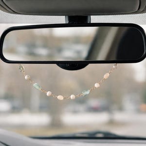 May include: A car rearview mirror with a decorative beaded strand hanging from it. The strand features white, light blue, and clear beads strung on a gold-colored chain. The background is blurred, suggesting a view through the windshield.
