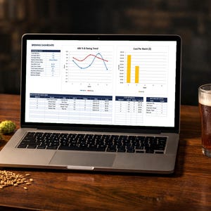 Op de afbeelding: Een laptop met een brouwdashboard met grafieken en gegevens staat op een houten tafel. Een glas bier, hop en granen staan ook op de tafel. Het scherm toont grafieken van ABV % & Rating Trend en Kosten per batch (€).