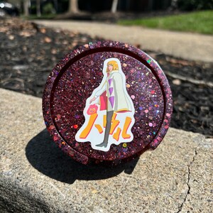 May include: A red glitter resin coaster featuring a character from the anime "Howl's Moving Castle" and the Japanese text "Howl" on it.