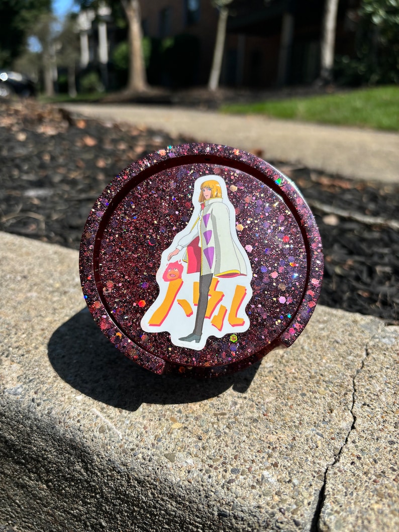 May include: A red glitter resin coaster featuring a character from the anime "Howl's Moving Castle" and the Japanese text "Howl" on it.
