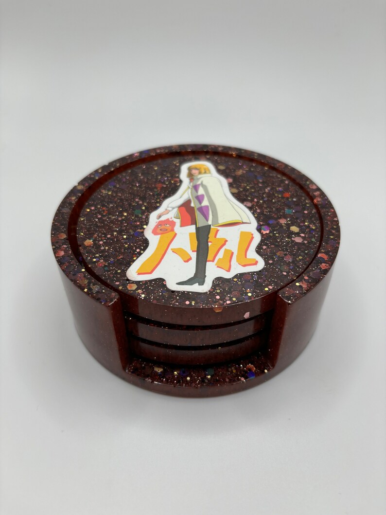 May include: Set of four brown coasters with a glittery finish. The top coaster features a printed image of a character from the anime "Howl's Moving Castle".