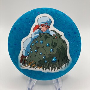 May include: A round, blue, glitter-filled resin coaster with a white sticker featuring a cartoon girl riding a green, multi-eyed creature.
