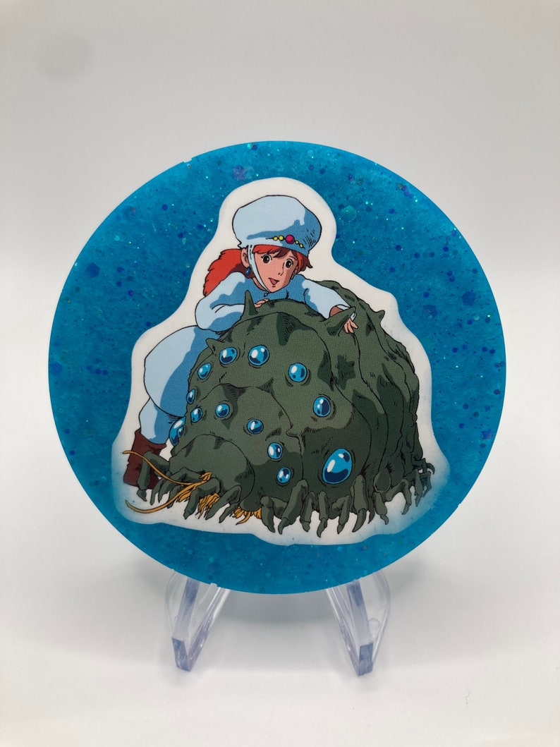 May include: A round, blue, glitter-filled resin coaster with a white sticker featuring a cartoon girl riding a green, multi-eyed creature.
