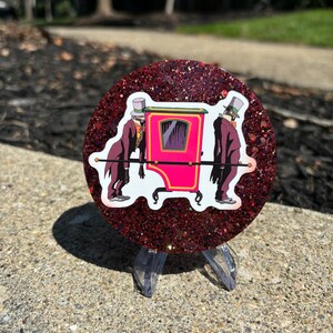 May include: A round, red glitter coaster with a white sticker of two figures in top hats carrying a pink sedan chair.