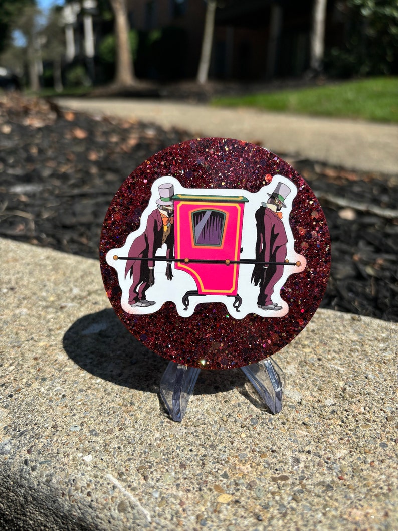 May include: A round, red glitter coaster with a white sticker of two figures in top hats carrying a pink sedan chair.