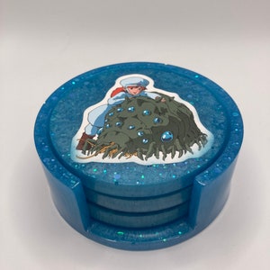 May include: A set of four blue coasters with a glittery finish. The top coaster features a printed image of a character from the anime film 'Castle in the Sky' riding a creature with multiple eyes.