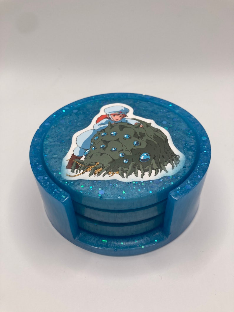 May include: A set of four blue coasters with a glittery finish. The top coaster features a printed image of a character from the anime film 'Castle in the Sky' riding a creature with multiple eyes.