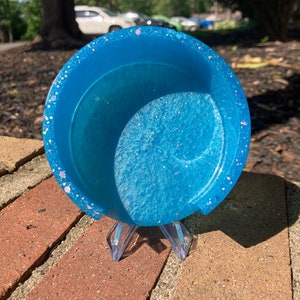 May include: A light blue resin coaster with iridescent glitter embedded in the resin. The coaster is sitting on a brick surface.