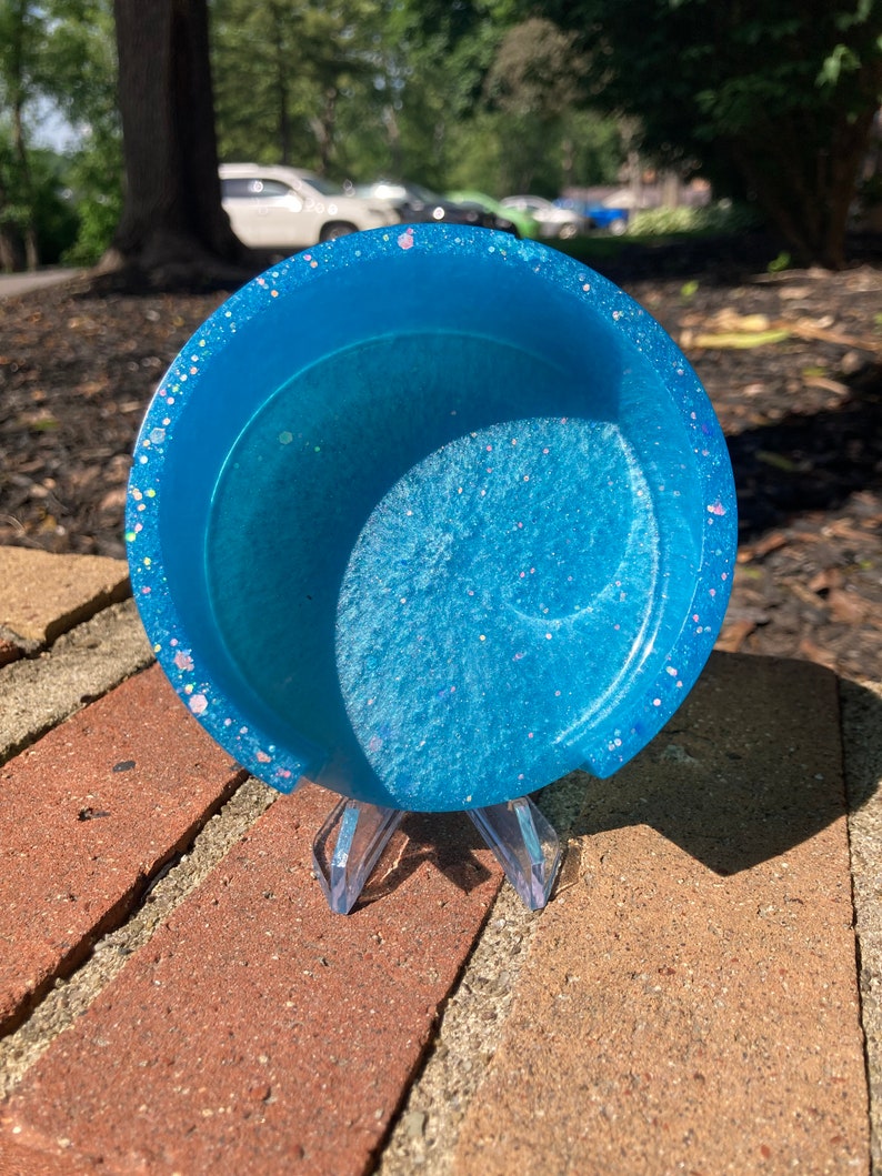 May include: A light blue resin coaster with iridescent glitter embedded in the resin. The coaster is sitting on a brick surface.