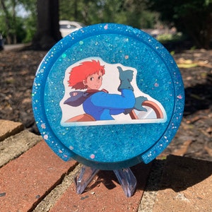 May include: A blue glitter resin coaster with a character from the anime film Castle in the Sky. The character is a young woman with red hair and blue eyes, wearing a blue shirt and a brown jacket. She is giving a thumbs up.