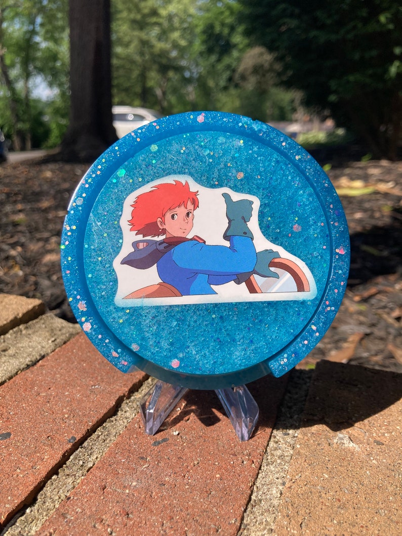 May include: A blue glitter resin coaster with a character from the anime film Castle in the Sky. The character is a young woman with red hair and blue eyes, wearing a blue shirt and a brown jacket. She is giving a thumbs up.