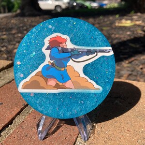 May include: A round blue and white glitter coaster with a printed image of a female character holding a rifle. The character is wearing a blue jumpsuit and has red hair.
