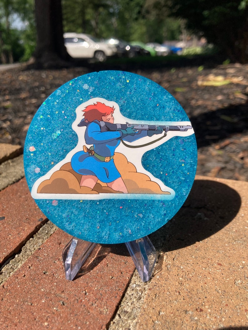 May include: A round blue and white glitter coaster with a printed image of a female character holding a rifle. The character is wearing a blue jumpsuit and has red hair.