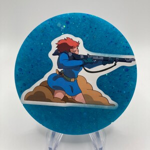 May include: A round blue glitter coaster with a white sticker featuring a cartoon woman with red hair holding a rifle. The woman is wearing a blue dress and is standing on a brown cloud.