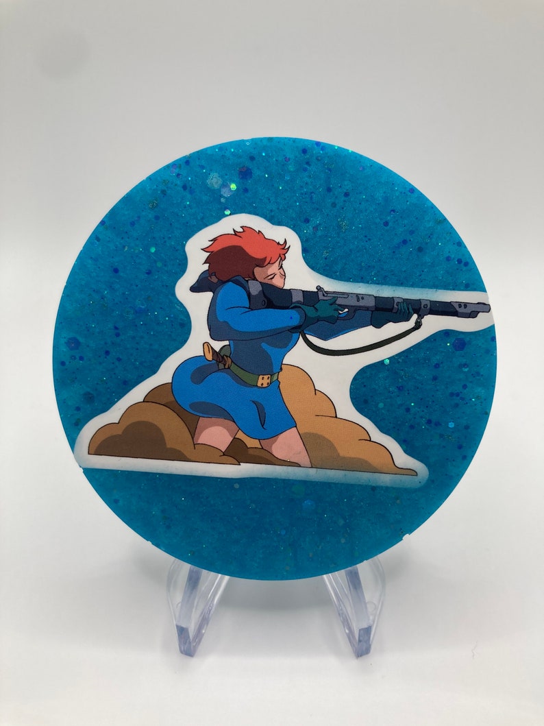 May include: A round blue glitter coaster with a white sticker featuring a cartoon woman with red hair holding a rifle. The woman is wearing a blue dress and is standing on a brown cloud.
