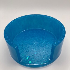 May include: A light blue round coaster with a white and blue glitter design. The coaster has a cut-out for a cup or mug.