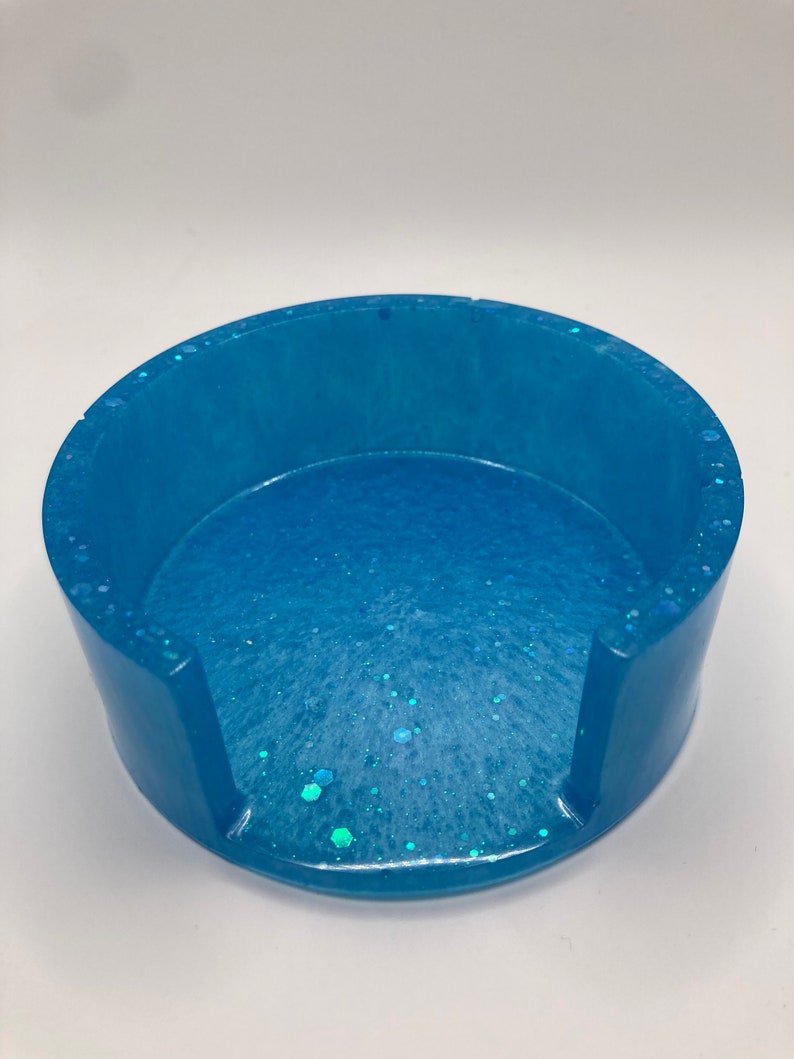 May include: A light blue round coaster with a white and blue glitter design. The coaster has a cut-out for a cup or mug.