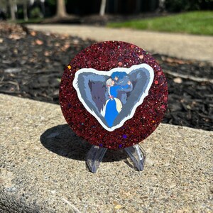 May include: A red glitter coaster featuring a sticker of a wolf and a woman embracing. The sticker is in the shape of a heart.