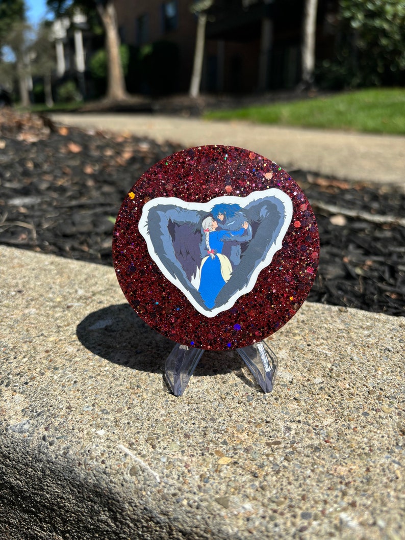 May include: A red glitter coaster featuring a sticker of a wolf and a woman embracing. The sticker is in the shape of a heart.