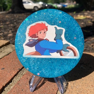 May include: A blue and white glitter resin coaster with a printed image of a cartoon character with red hair giving a thumbs up.
