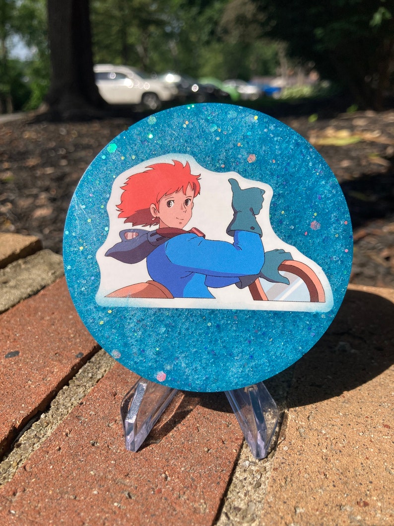 May include: A blue and white glitter resin coaster with a printed image of a cartoon character with red hair giving a thumbs up.