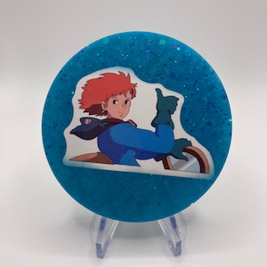 May include: A round blue glitter coaster with a white sticker featuring an anime character giving a thumbs up.