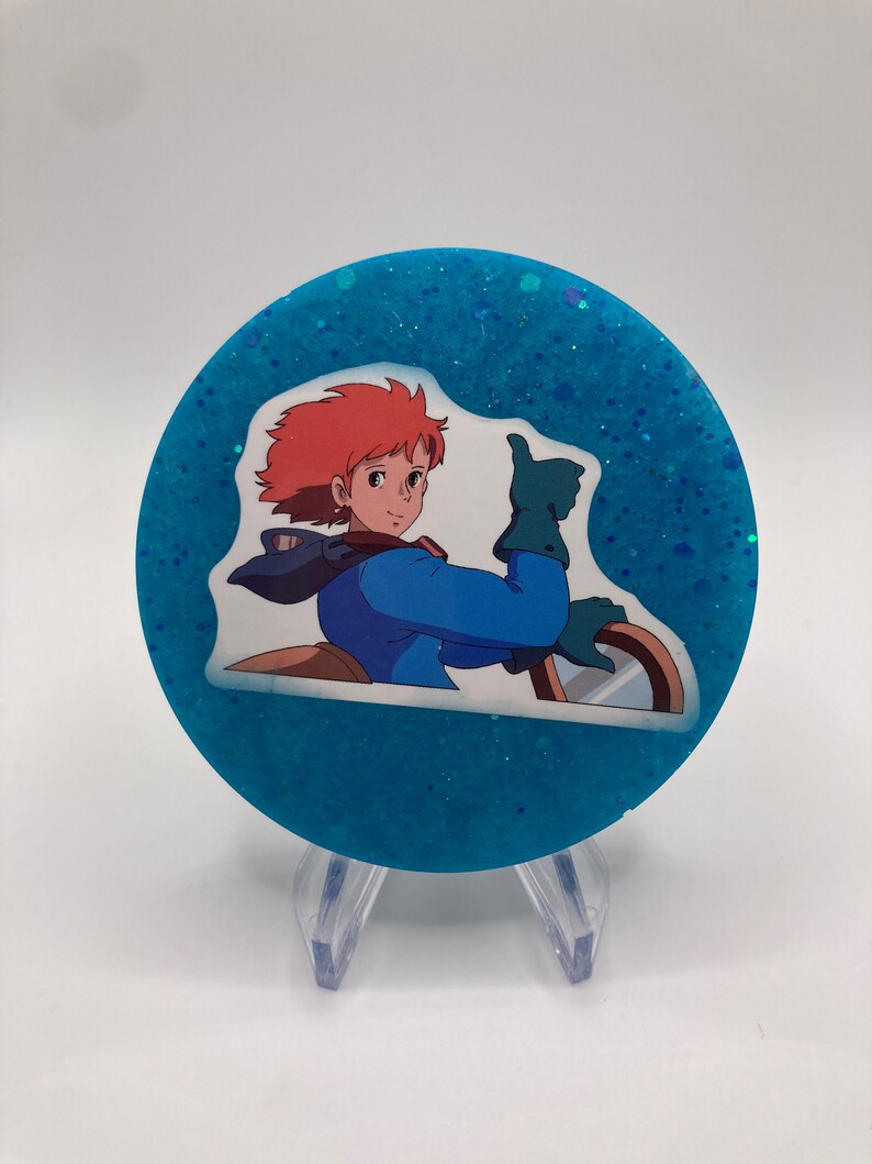 May include: A round blue glitter coaster with a white sticker featuring an anime character giving a thumbs up.