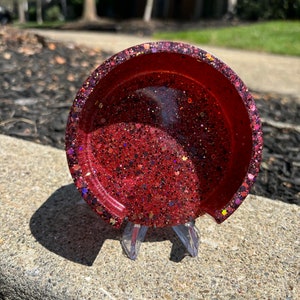 May include: A red glitter resin coaster with a clear resin base. The coaster is on a clear plastic stand.