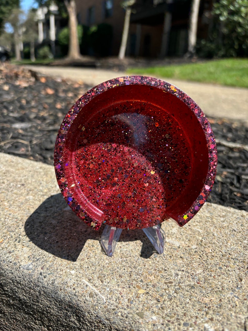 May include: A red glitter resin coaster with a clear resin base. The coaster is on a clear plastic stand.