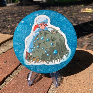May include: A round blue resin coaster with glitter embedded in the resin. The coaster features a printed image of a girl riding a large, multi-eyed, green creature with a long, yellow beard.