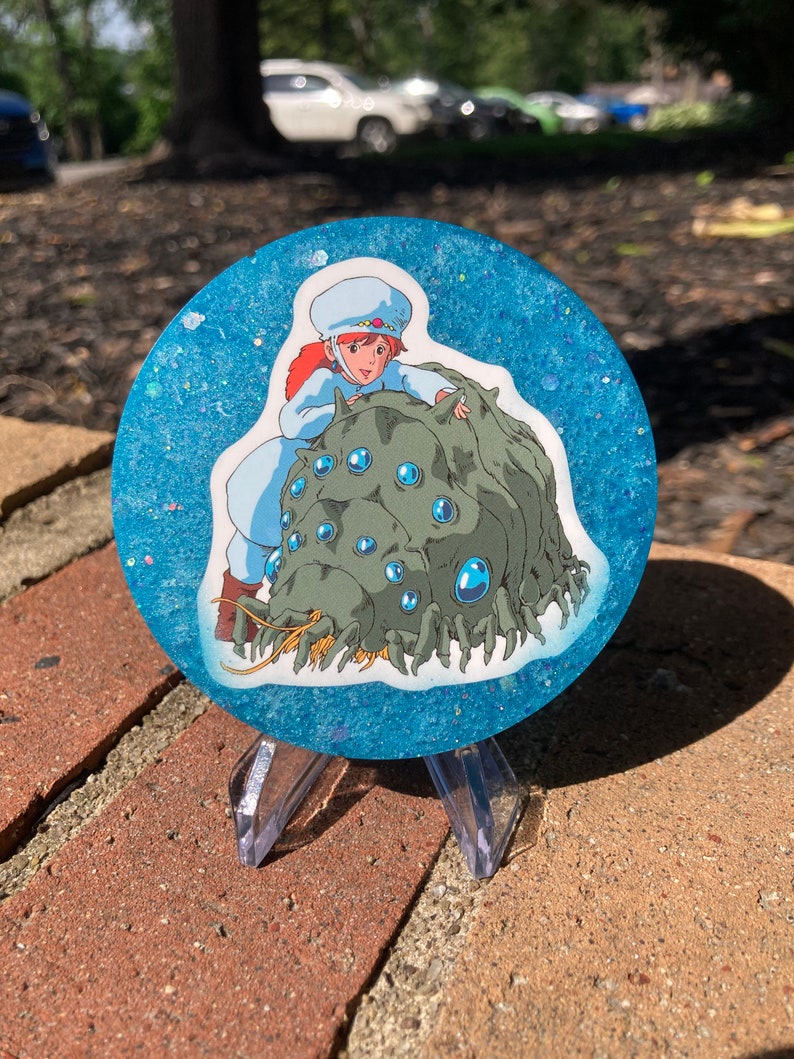 May include: A round blue resin coaster with glitter embedded in the resin. The coaster features a printed image of a girl riding a large, multi-eyed, green creature with a long, yellow beard.