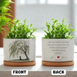 May include: Two white ceramic planters with green plants, each on a wooden saucer. One planter features a willow tree illustration with a couple. The other has a quote and names with a heart symbol.