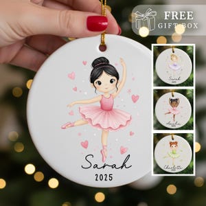 May include: A white ceramic ornament featuring a ballerina in a pink tutu, with the name "Sarah" and the year "2025" written below. The ornament has a gold string for hanging and includes a "FREE GIFT BOX" graphic.