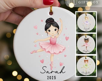 May include: A white ceramic ornament featuring a ballerina in a pink tutu, with the name "Sarah" and the year "2025" written below. The ornament has a gold string for hanging and includes a "FREE GIFT BOX" graphic.
