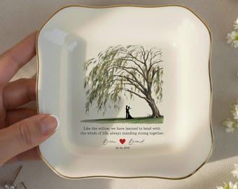 May include: A square, off-white ceramic trinket dish with a gold rim. The center features a willow tree illustration with two figures. Text reads, "Like the willow, we have learned to bend with the winds of life, always standing strong together."