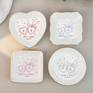 May include: Four white ceramic ring dishes with gold rims. Each dish features a heart-shaped design with two connected hearts and personalized text. The shapes include a heart, square, and round dish. The text includes names and dates.