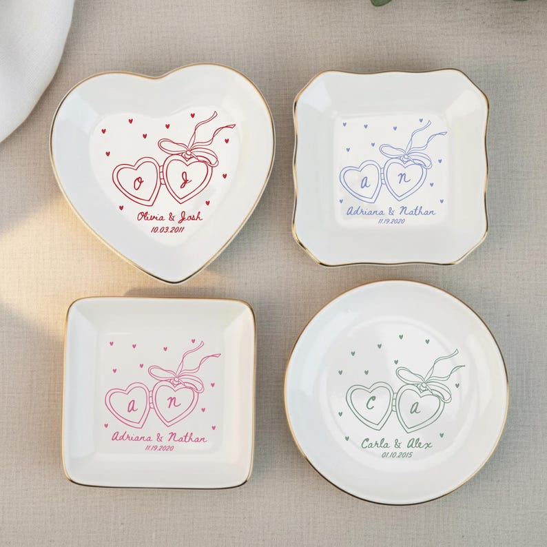 May include: Four white ceramic ring dishes with gold rims. Each dish features a heart-shaped design with two connected hearts and personalized text. The shapes include a heart, square, and round dish. The text includes names and dates.