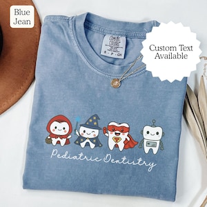 K&ouml;nnte beinhalten: Ein blaues Comfort Colors T-Shirt mit vier Zeichentrickz&auml;hnen, die als M&auml;rchenfigur, Zauberer, Superheld und Roboter verkleidet sind. Die Worte "Pediatric Dentistry" sind darunter geschrieben. Eine goldene Halskette ist sichtbar.