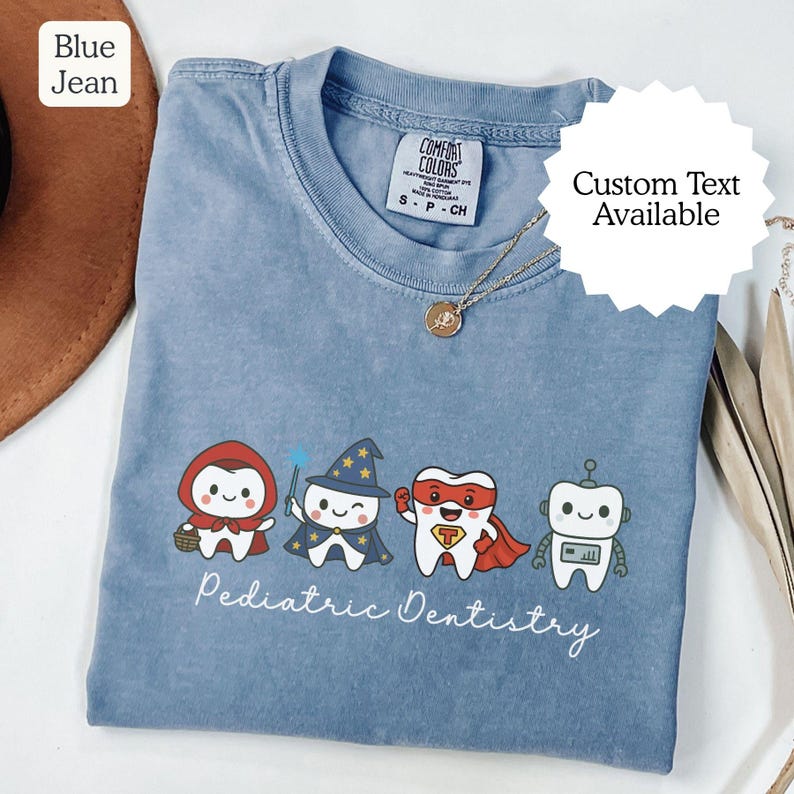 K&ouml;nnte beinhalten: Ein blaues Comfort Colors T-Shirt mit vier Zeichentrickz&auml;hnen, die als M&auml;rchenfigur, Zauberer, Superheld und Roboter verkleidet sind. Die Worte "Pediatric Dentistry" sind darunter geschrieben. Eine goldene Halskette ist sichtbar.