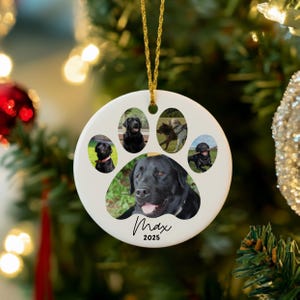 May include: White ceramic Christmas ornament shaped like a dog paw print, featuring photos of dogs. The ornament has a gold string for hanging and includes the name "Max" and the year "2025" in a script font.
