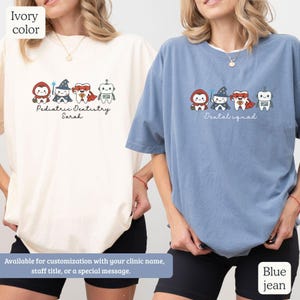 K&ouml;nnte beinhalten: Zwei T-Shirts, eines in Elfenbein und eines in Jeansblau, mit einem Cartoon-Design einer Zahnarztgruppe. Das elfenbeinfarbene Shirt tr&auml;gt den Text "Pediatric Dentistry Sarah", w&auml;hrend das Jeansblau-Shirt "Dental squad" sagt. Beide Shirts sind anpassbar.
