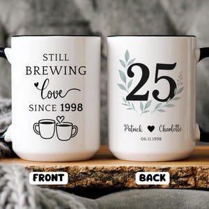 Pu&ograve; includere: Tazza in ceramica bianca con manico e bordo neri. La parte anteriore della tazza recita "Still Brewing Love Since 1998" con illustrazioni di tazze da caff&egrave;. Il retro della tazza mostra il numero "25" con nomi e una data.
